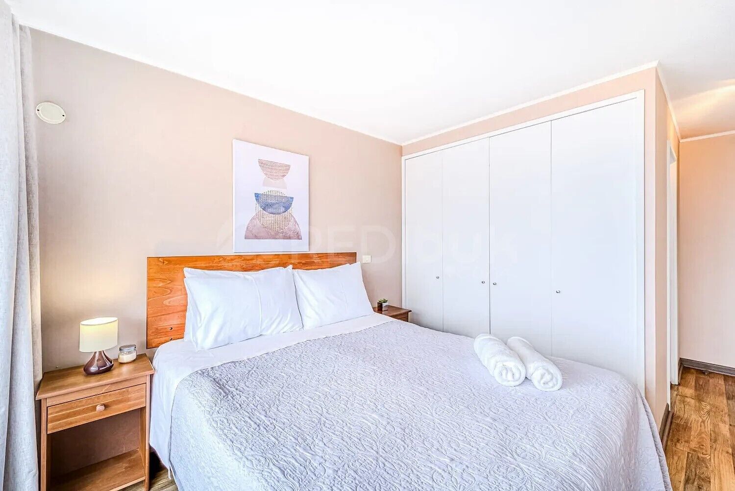 Una cama bien hecha con sábanas blancas, un cabecero de madera y una mesita de noche con una lámpara.