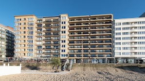 Ein Strandhotel mit mehreren Balkonen, ein Sandstrand im Vordergrund und ein klarer, blauer Himmel.