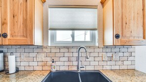 Cozinha com bancada de granito, pia dupla, armários de madeira, janela com persiana e painel de backsplash de tijolos.