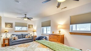 Quarto com cama, sofá, cômoda, ventilador de teto e duas janelas com persianas.
