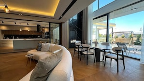 Un salon moderne comprenant un canapé, une table à manger et des chaises. On y trouve également un coin cuisine avec un bar et une grande fenêtre donnant sur l’extérieur.