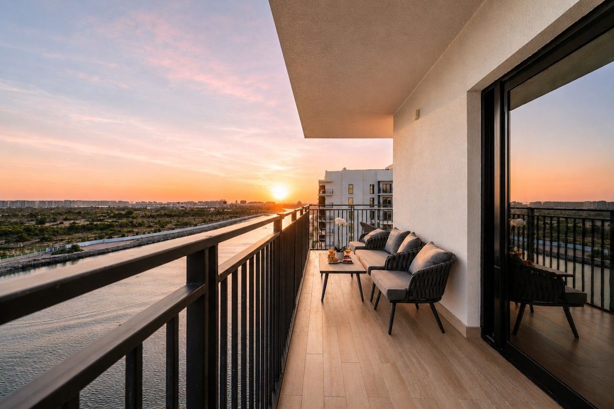 Un balcón con sofá y mesa, con vista a un río y al paisaje urbano al atardecer.