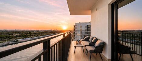 A balcony with a sofa and table, overlooking a river and cityscape during sunset.