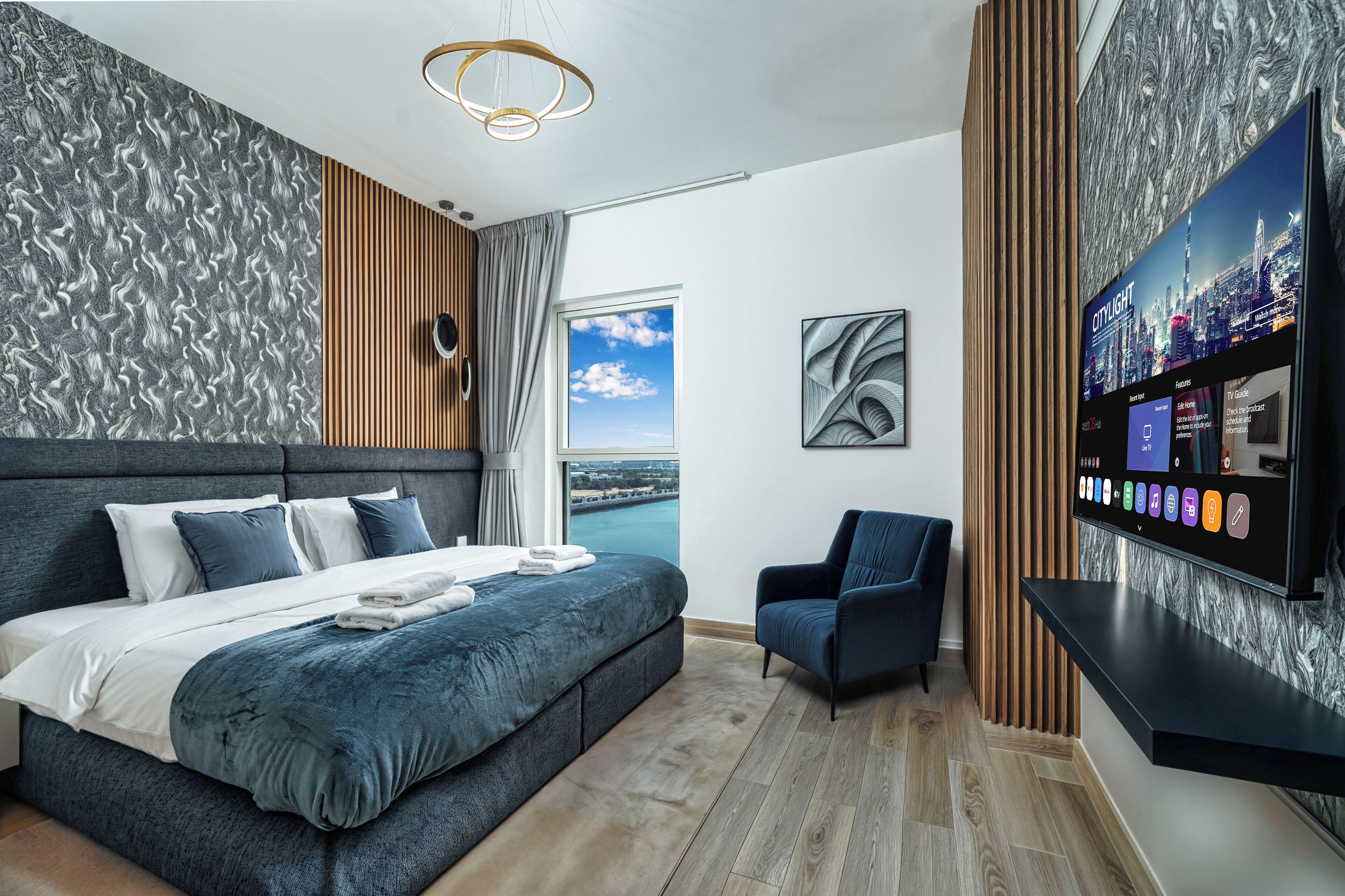 Ein modernes Schlafzimmer mit einem großen Bett, einem Fernsehgerät mit Stadtansicht und Blick auf das Wasser.