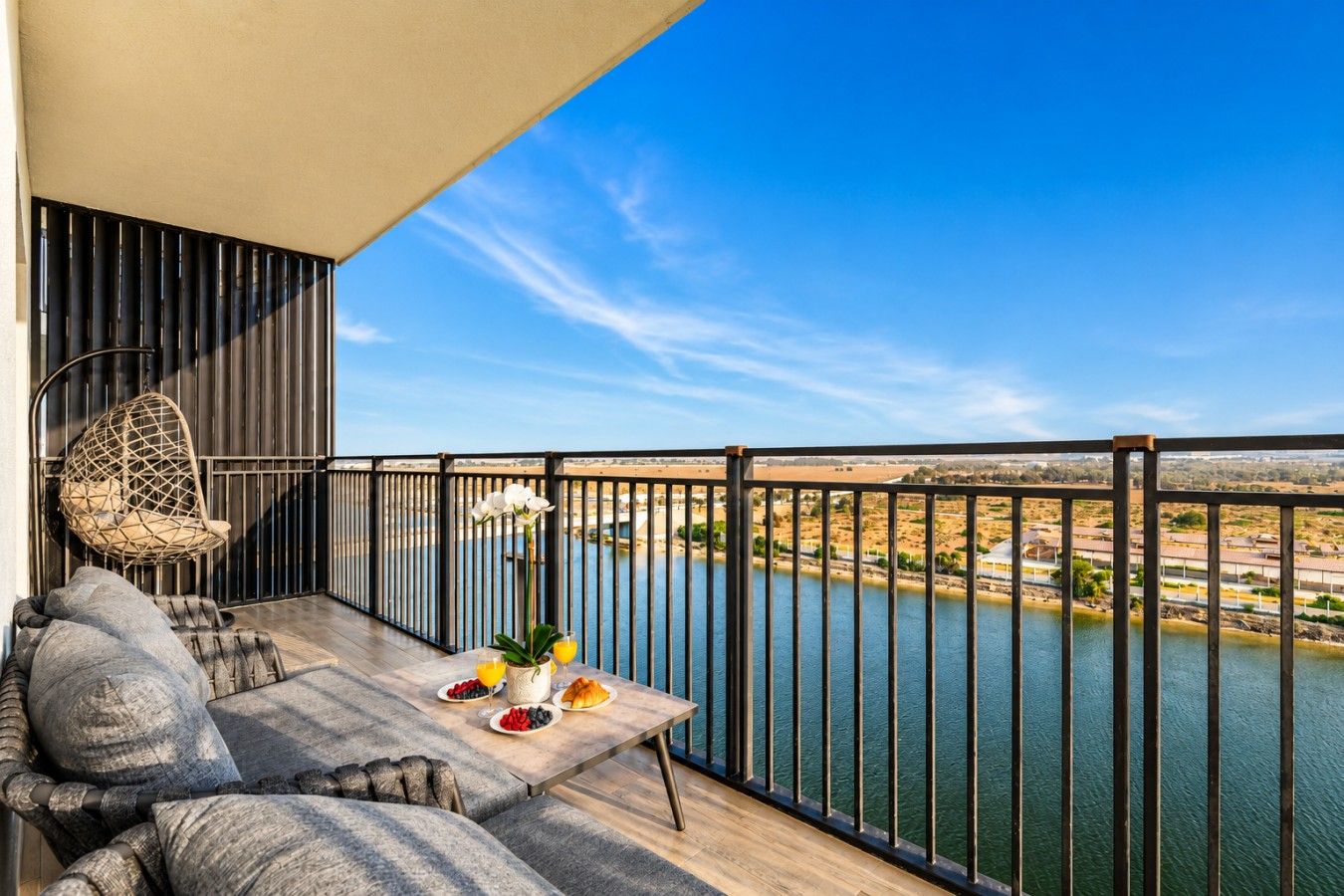 A balcony with a view of a body of water, a hanging chair, a table with fruit and a vase, and a railing.