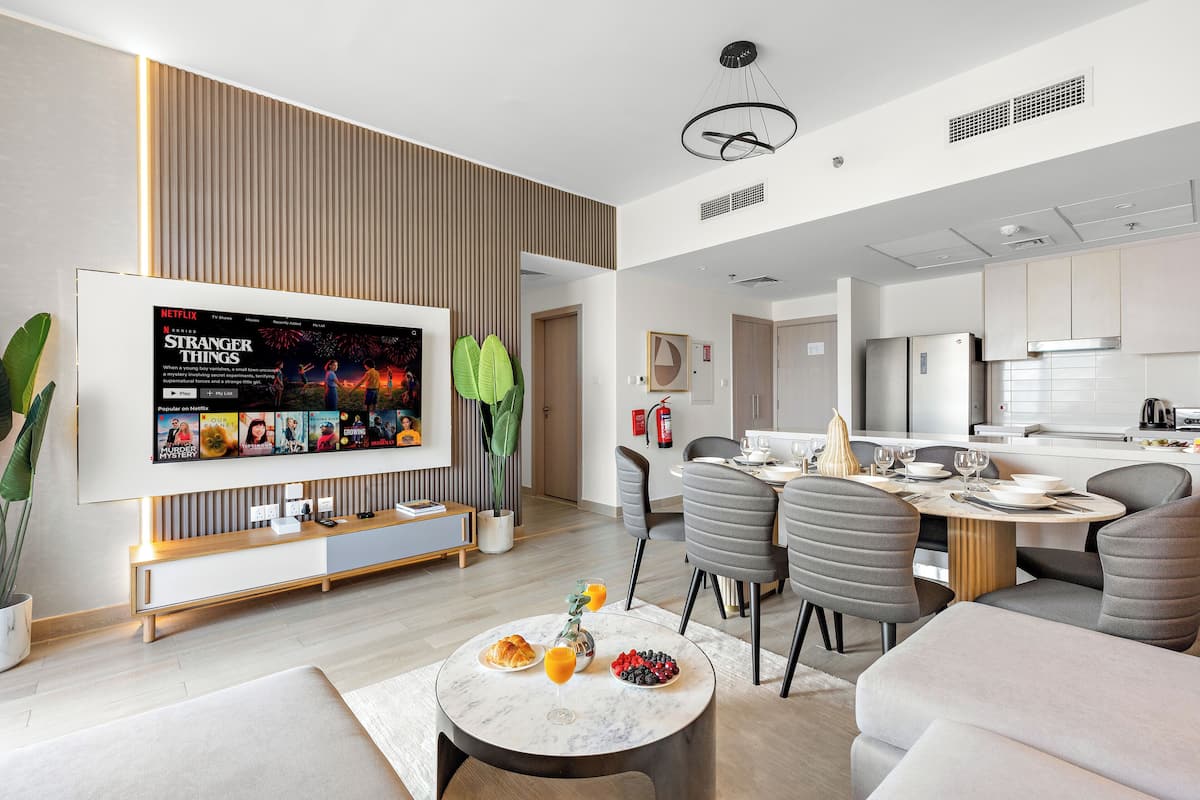 A modern living room with a flat-screen TV displaying 'Stranger Things', a dining area with a table set for two, and a kitchen in the background.