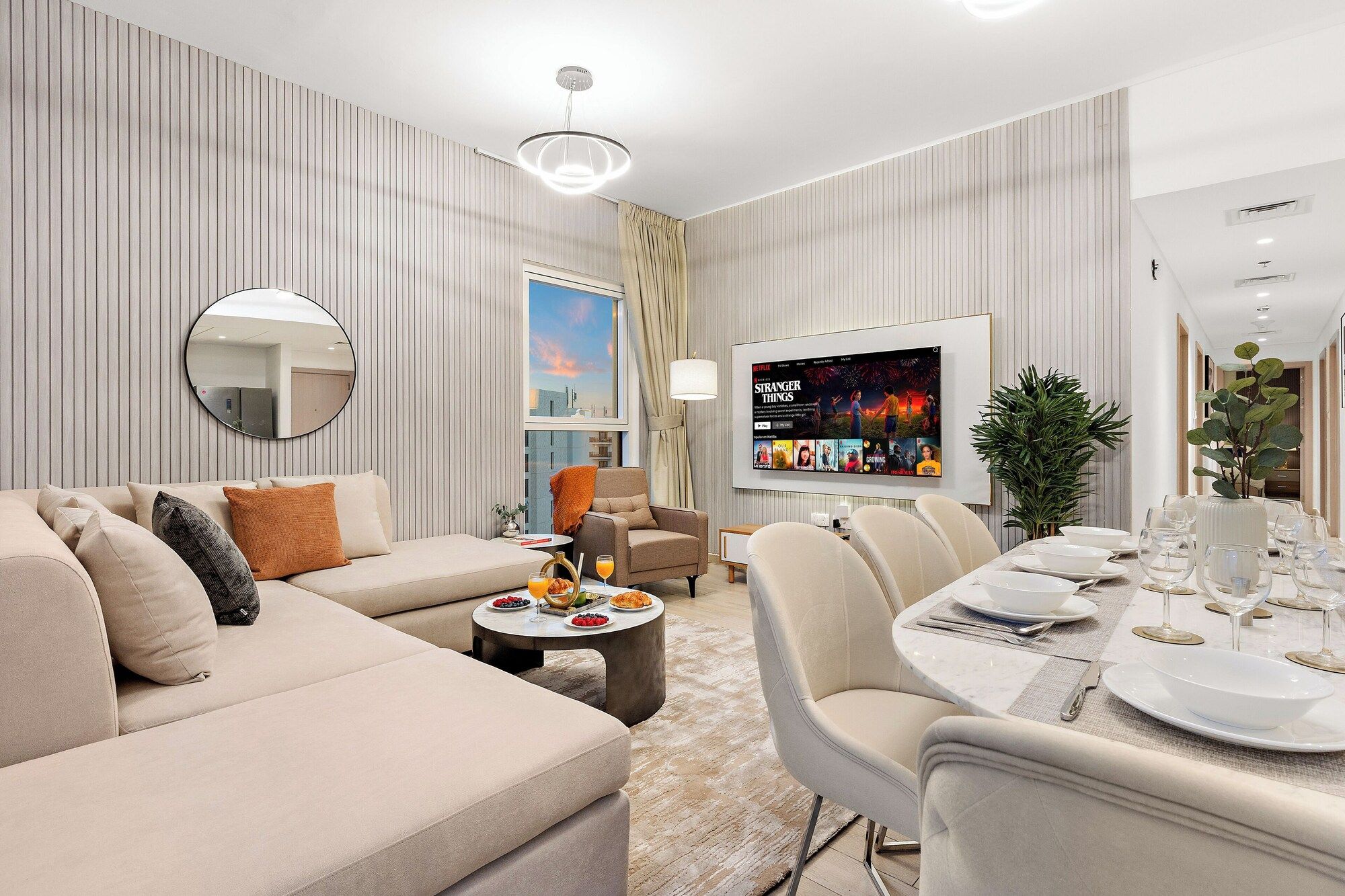 Un salon moderne comprenant un canapé, des fauteuils, une table à manger pour quatre personnes, un téléviseur fixé au mur et une grande fenêtre avec des rideaux.