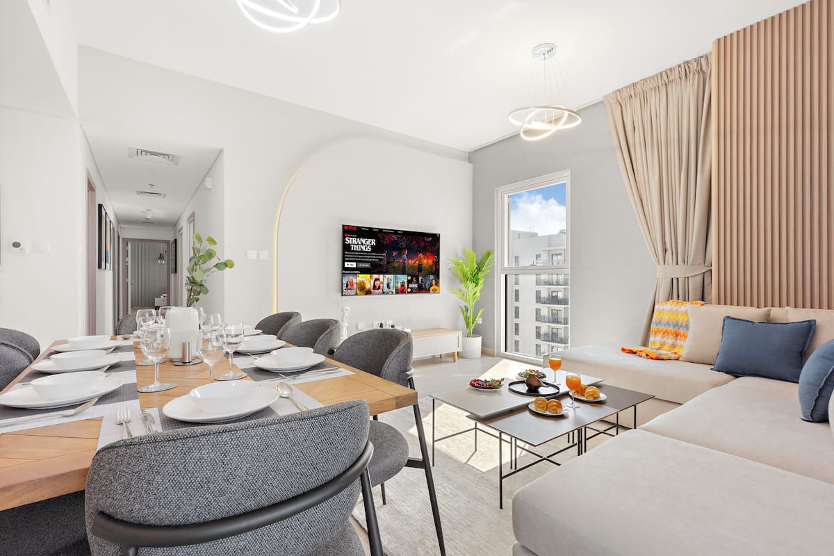 A modern living room with a dining area, a TV mounted on the wall, and a large window with curtains.