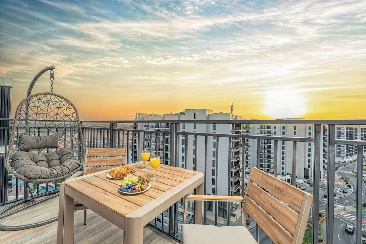 A balcony with a wooden table, a hanging chair, and a view of the sunset over a cityscape.