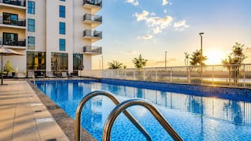 Una piscina con bordo piastrellato, scala in metallo e un edificio con balconi sullo sfondo.