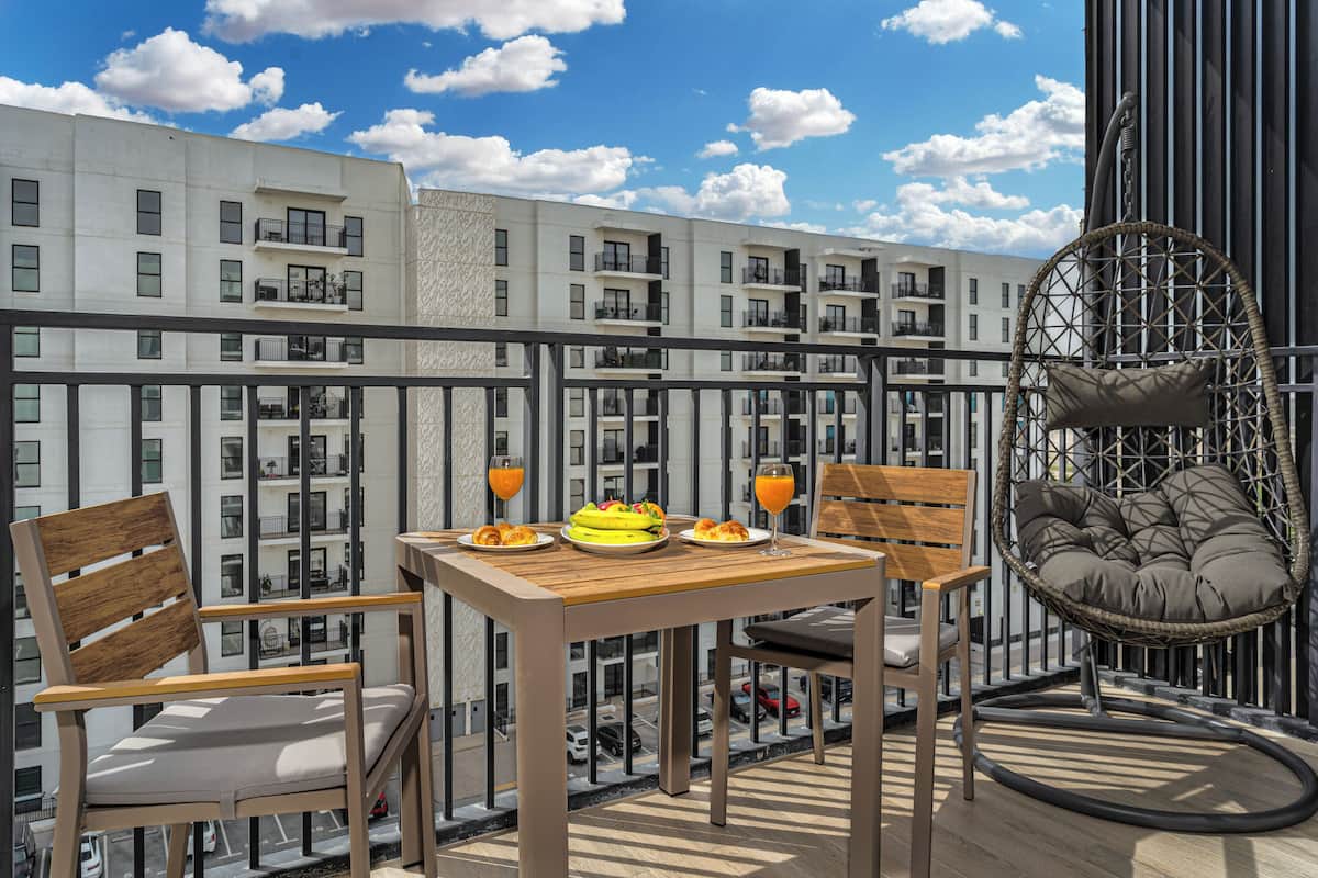 A balcony with a wooden table set for breakfast, two chairs, and a wicker chair with cushions.