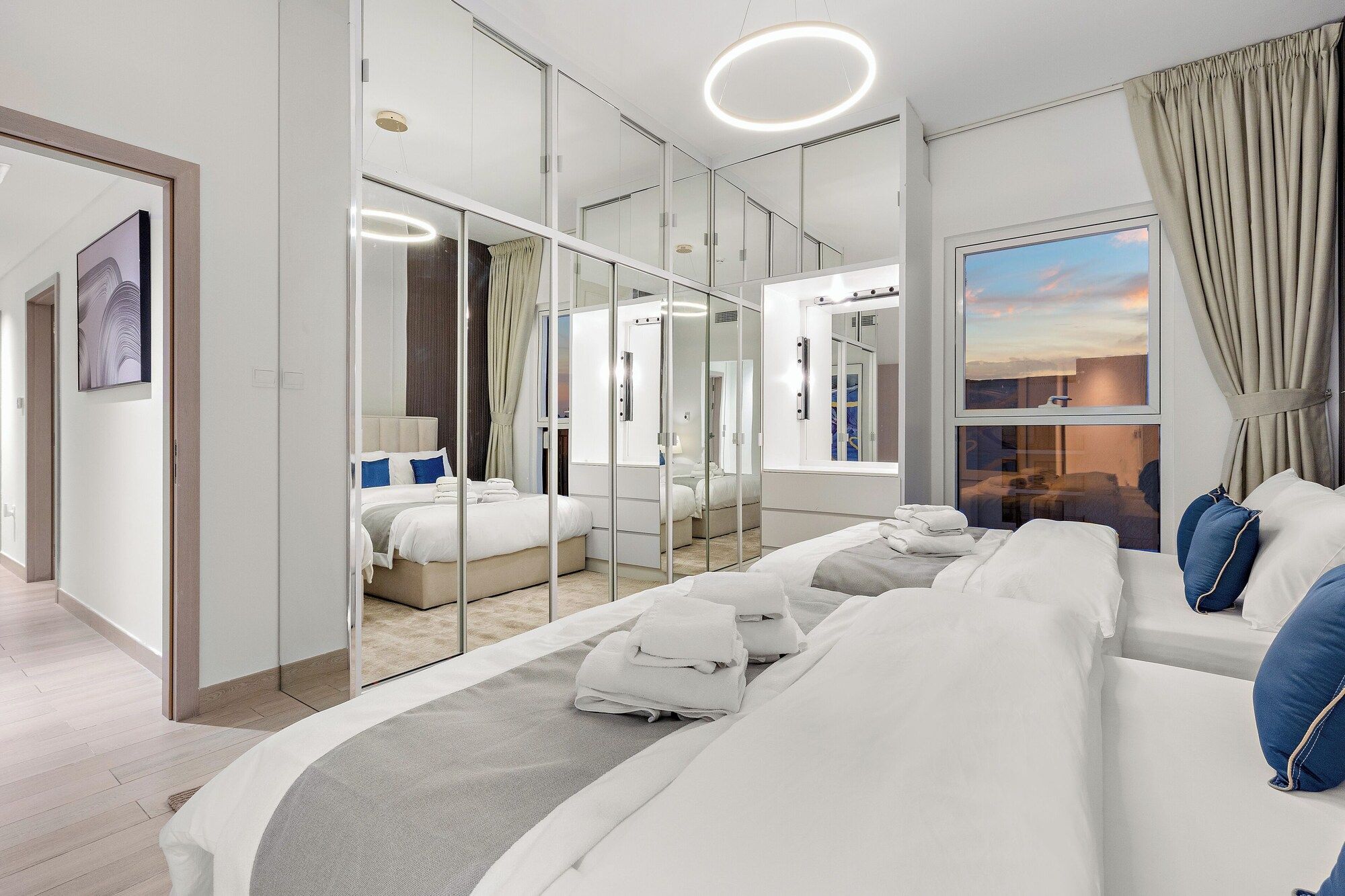 A modern hotel room with a large bed, a walk-in closet, a large mirror, and a view of the sunset.