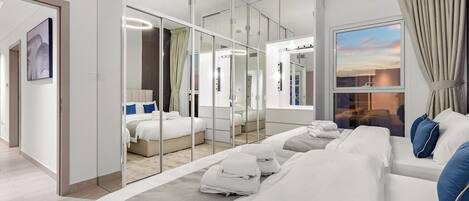 A modern hotel room with a large bed, a walk-in closet, a large mirror, and a view of the sunset.
