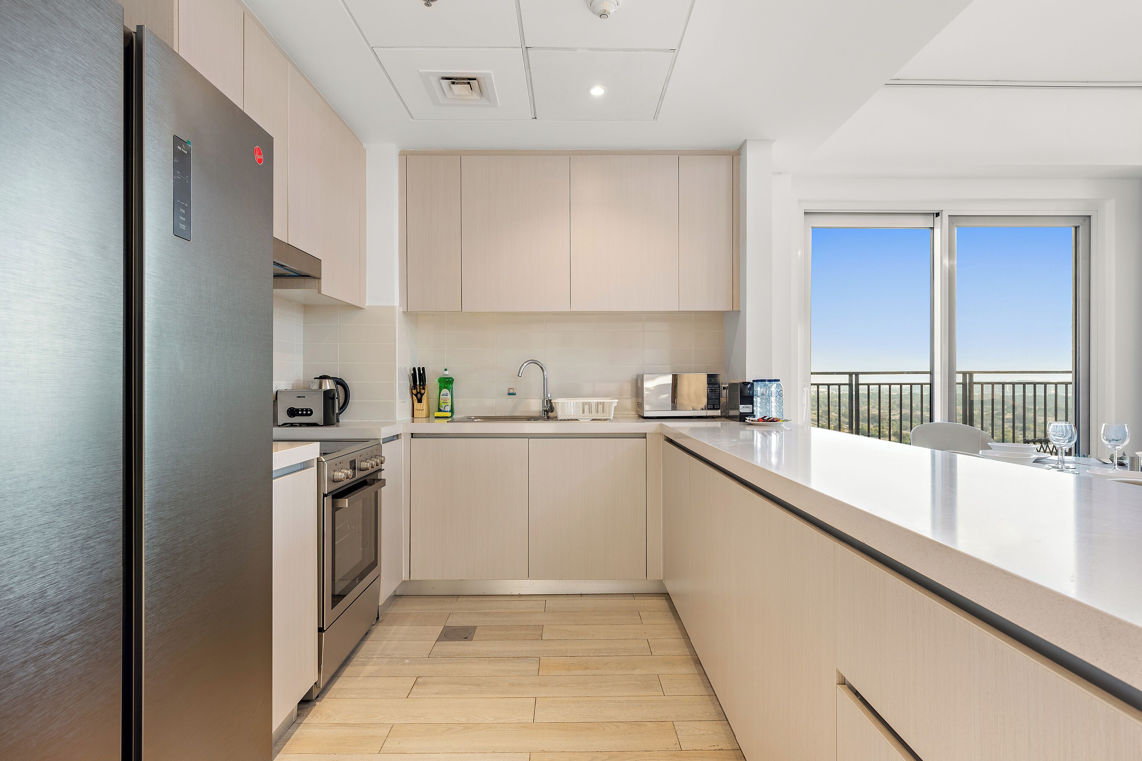 A modern kitchen with stainless steel appliances, light wood cabinetry, and a large window offering a view of the outdoors.