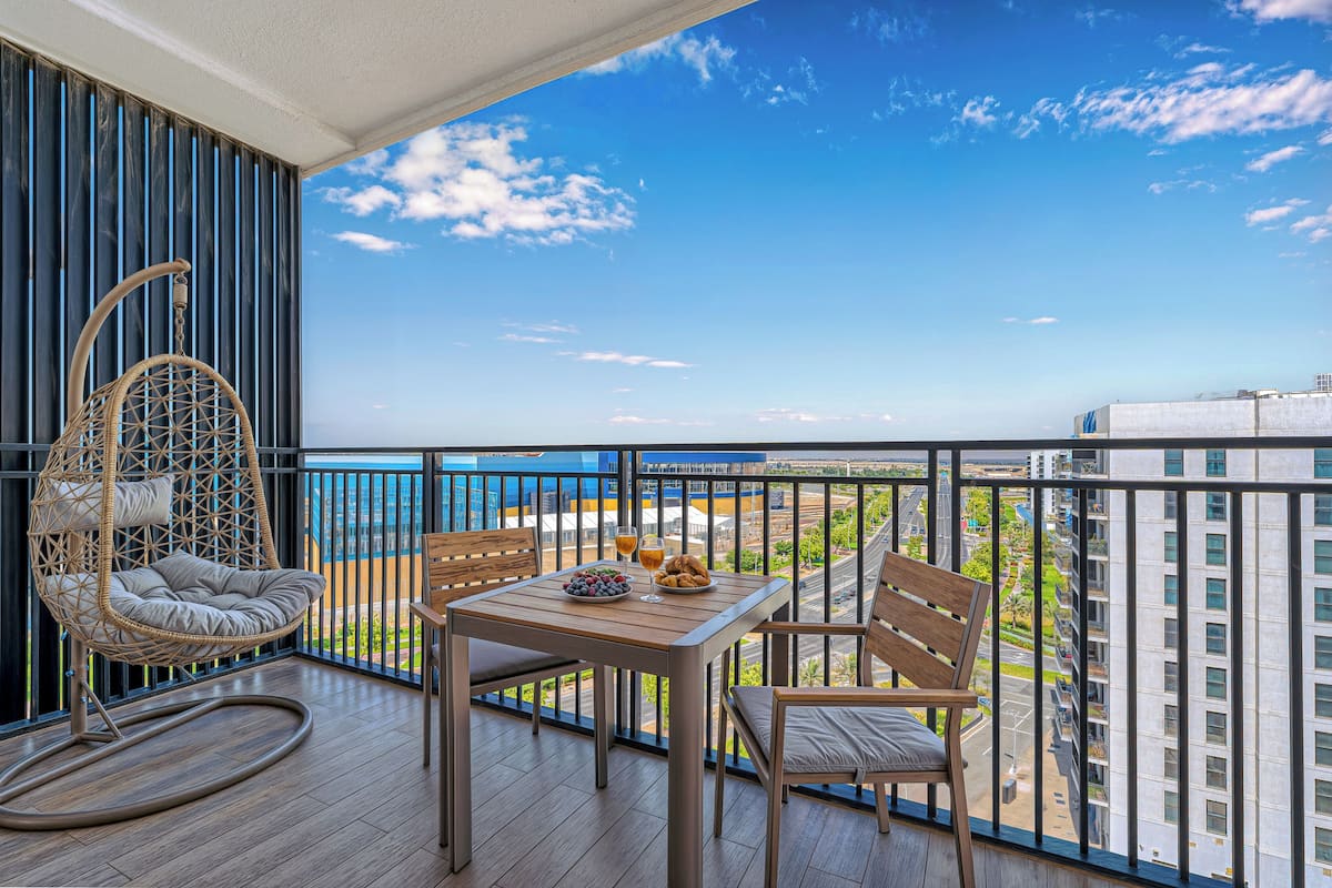 A balcony with a wicker chair, a wooden table with a bowl of fruit and glasses, and a view of the city and sea.