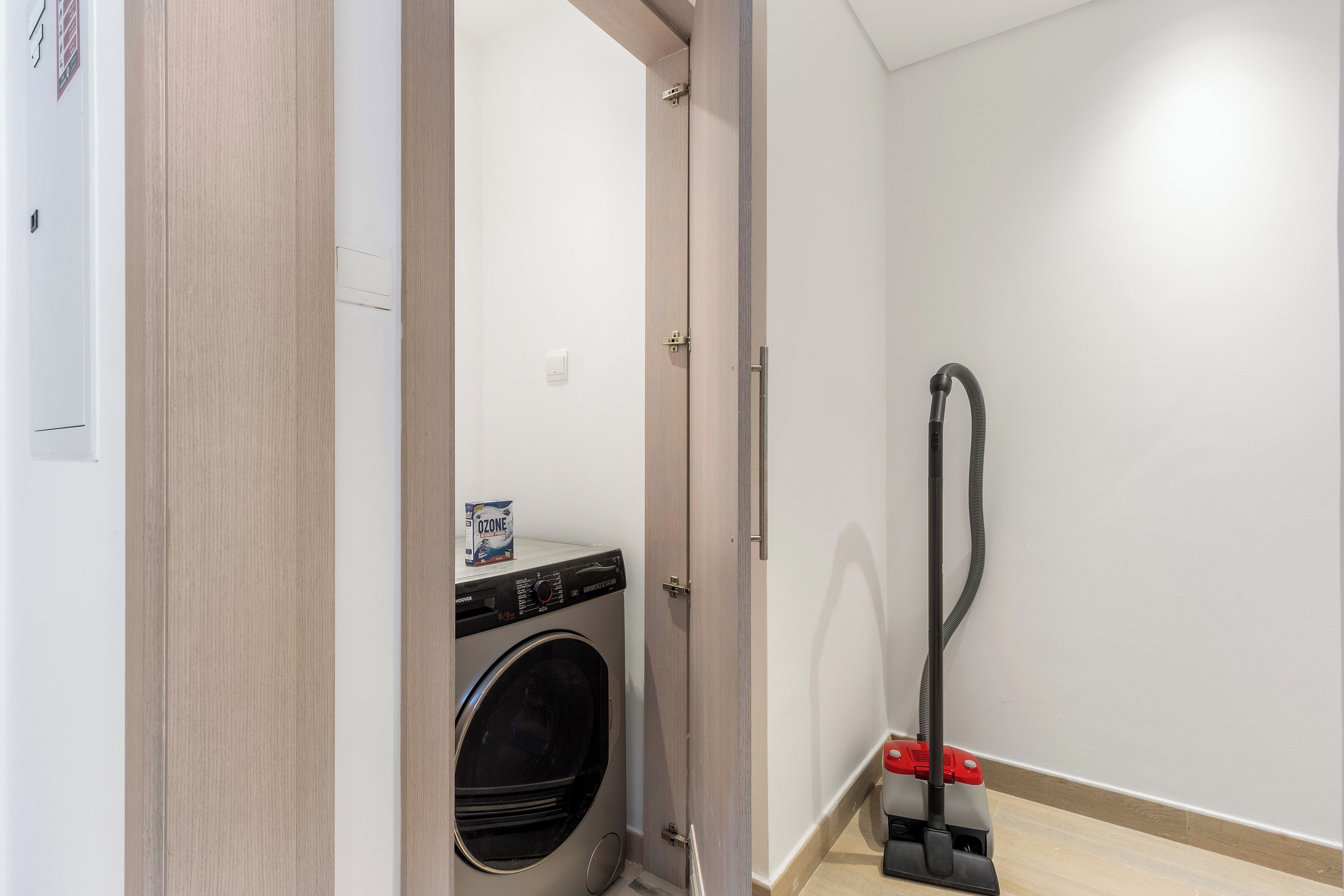 A laundry area with a washing machine, a vacuum cleaner, and a door leading to another room.