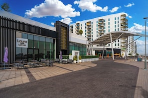 Un hotel moderno con un área de estar al aire libre cubierta, una entrada amplia y un edificio de varios pisos al fondo.