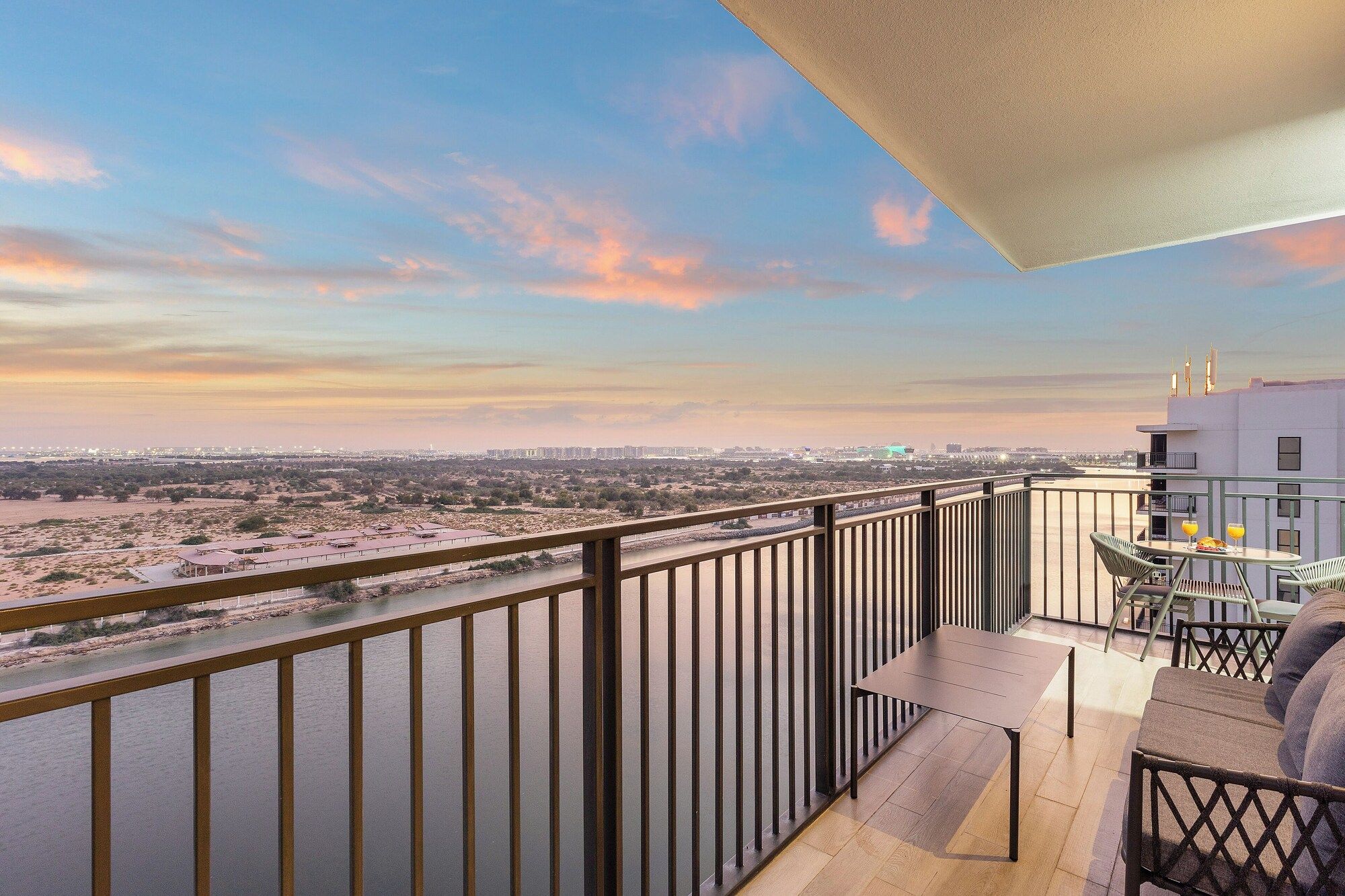 A balcony with a view of a river and distant cityscape, a table with chairs, and a railing.
