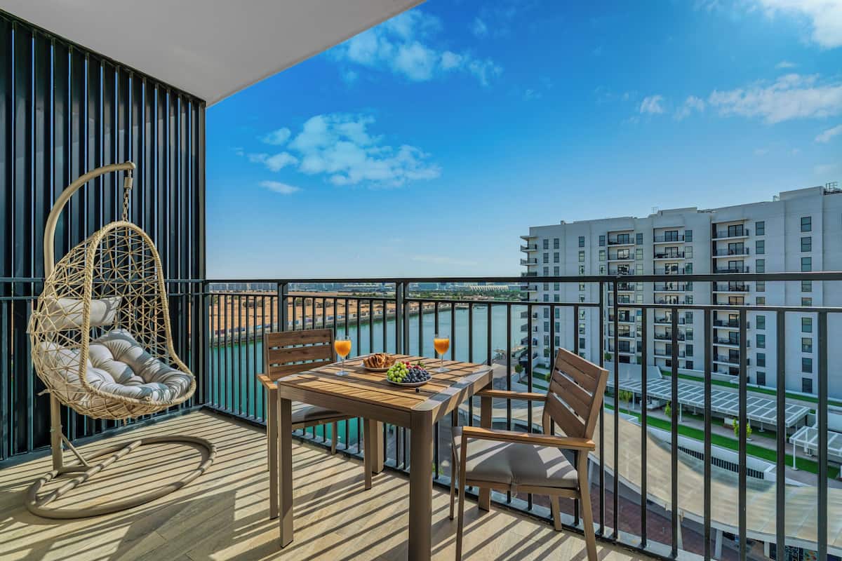 A balcony with a wicker chair, wooden table, and two chairs. There is a view of a building and a body of water in the background.