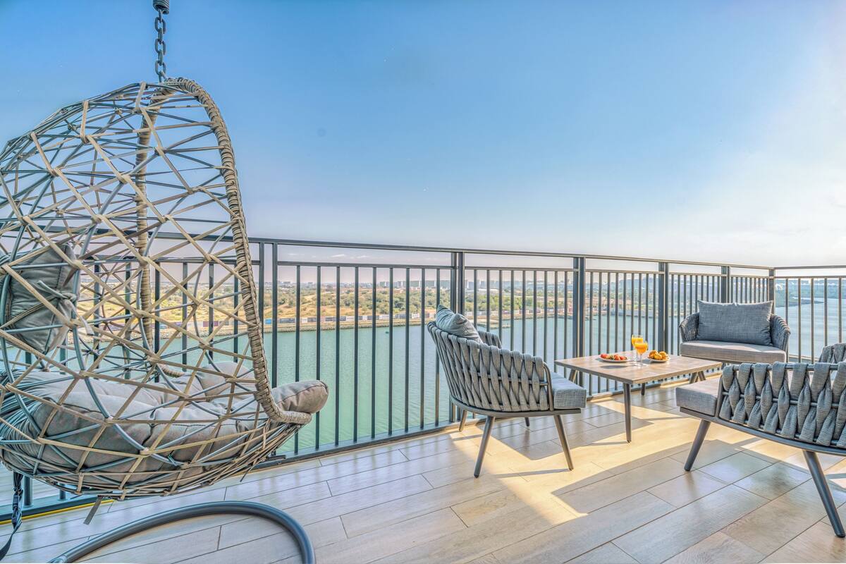 A balcony with a hanging chair, two armchairs, a small table, and a view of a body of water.