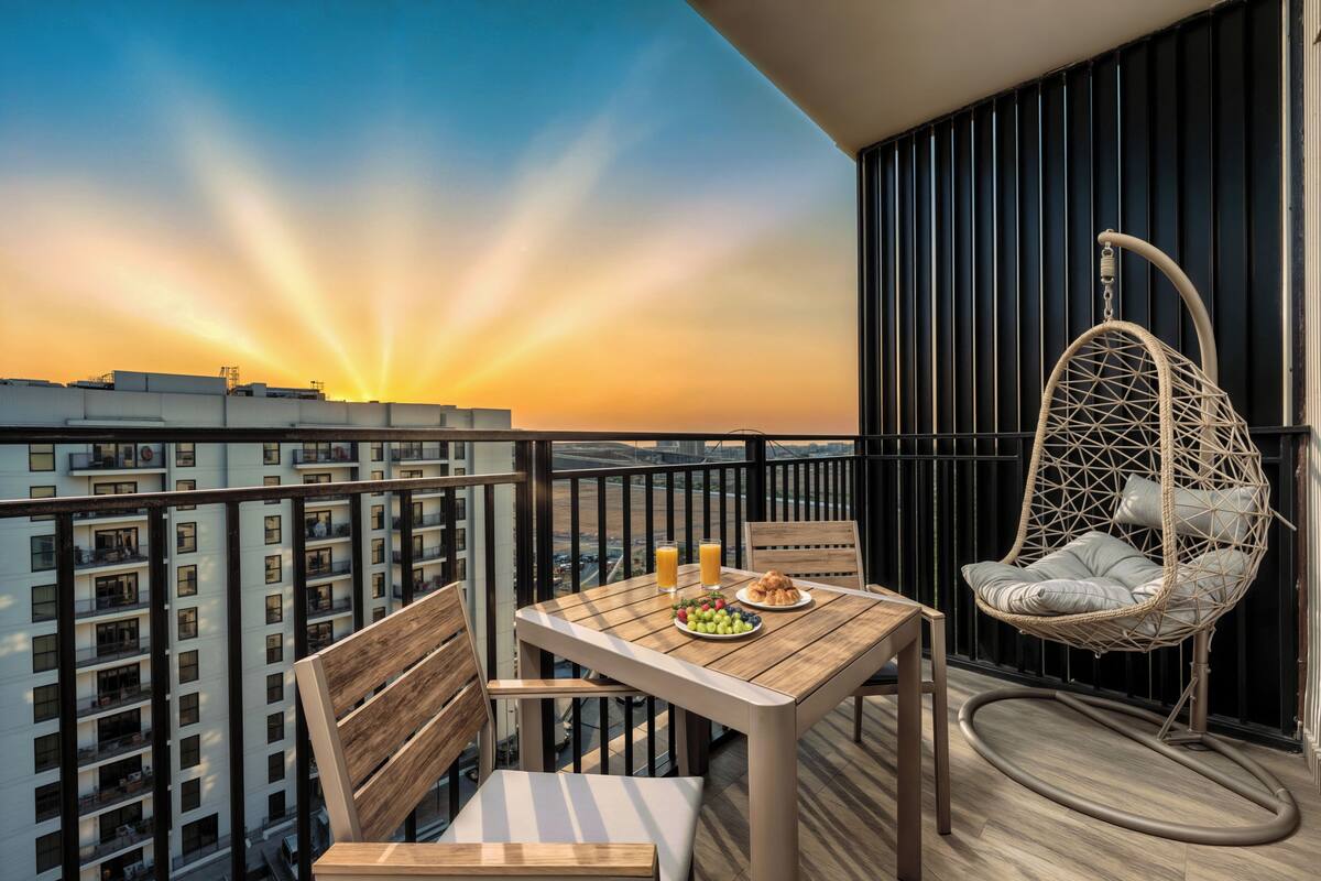 A balcony with a table set for breakfast, a wicker chair, and a view of a cityscape at sunset.