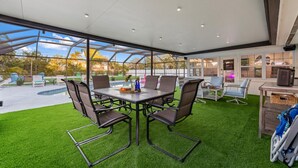 A covered outdoor area with a table and chairs, artificial grass, and a view of a swimming pool and trees.
