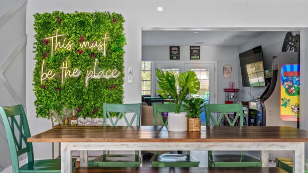 A dining area with a wooden table, green chairs, and a wall decorated with greenery and a neon sign.