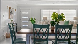 A modern living room with a dining area, featuring a large window with blinds, a sofa, two chairs, and decorative plants.