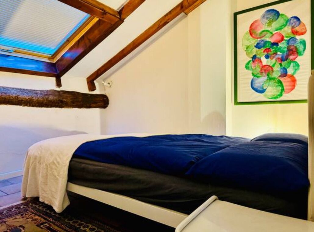A bedroom with a skylight, a bed with a blue and white duvet, a wooden beam, and a colorful abstract painting on the wall.
