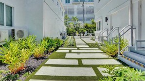 A narrow pathway with a checkered pattern leading to a building entrance, flanked by greenery and air conditioning units. - Walk to FL Beach | Chic Studio Suite + Pool (Fort Lauderdale)