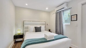 A modern hotel bedroom with a large bed, a bedside table with a lamp, and a window with curtains. - Walk to FL Beach | Chic Studio Suite + Pool (Fort Lauderdale)