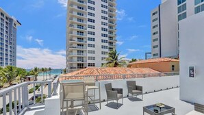 Una terraza en la azotea con mobiliario exterior, vista a la playa y edificios altos al fondo.