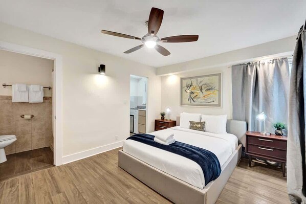 A hotel room with a bed, a ceiling fan, a nightstand, a lamp, a painting on the wall, and a bathroom with a toilet and towel rack. - Eden Studio Suite Walk to FLT Beach (Fort Lauderdale)