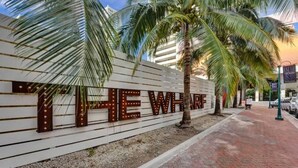 Ein Schild mit beleuchteten Buchstaben, die "THE WHARF" schreiben, an einer weißen Wand, mit Palmen und einem Pflasterweg.