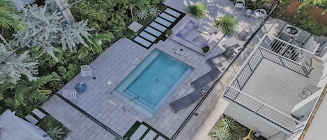 An aerial view of a rooftop pool area with a rectangular pool, lounge chairs, and surrounding greenery.