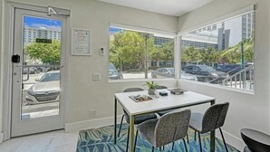 Une pièce comprenant une table à manger, des chaises, une porte et de grandes fenêtres donnant sur des voitures garées et des immeubles.
