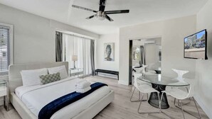 Una camera d'albergo moderna con un letto grande, un tavolo da pranzo con sedie, una ventola a soffitto e un'ampia finestra con tende.