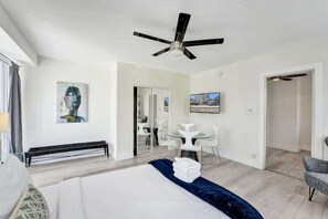 Une chambre d’hôtel moderne, équipée d’un lit, d’un ventilateur de plafond, d’un tableau, d’un coin repas avec des chaises et offrant une vue à travers une porte vitrée.