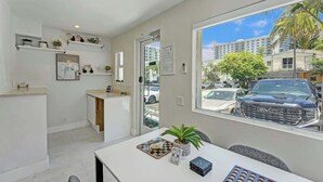 A modern kitchen with white cabinetry, a dining area with a table, and a large window offering a view of a parking lot and buildings.