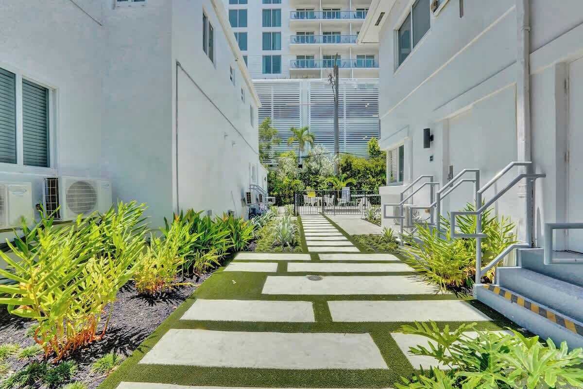A narrow pathway with stepping stones leading to a building entrance, flanked by greenery and air conditioning units.