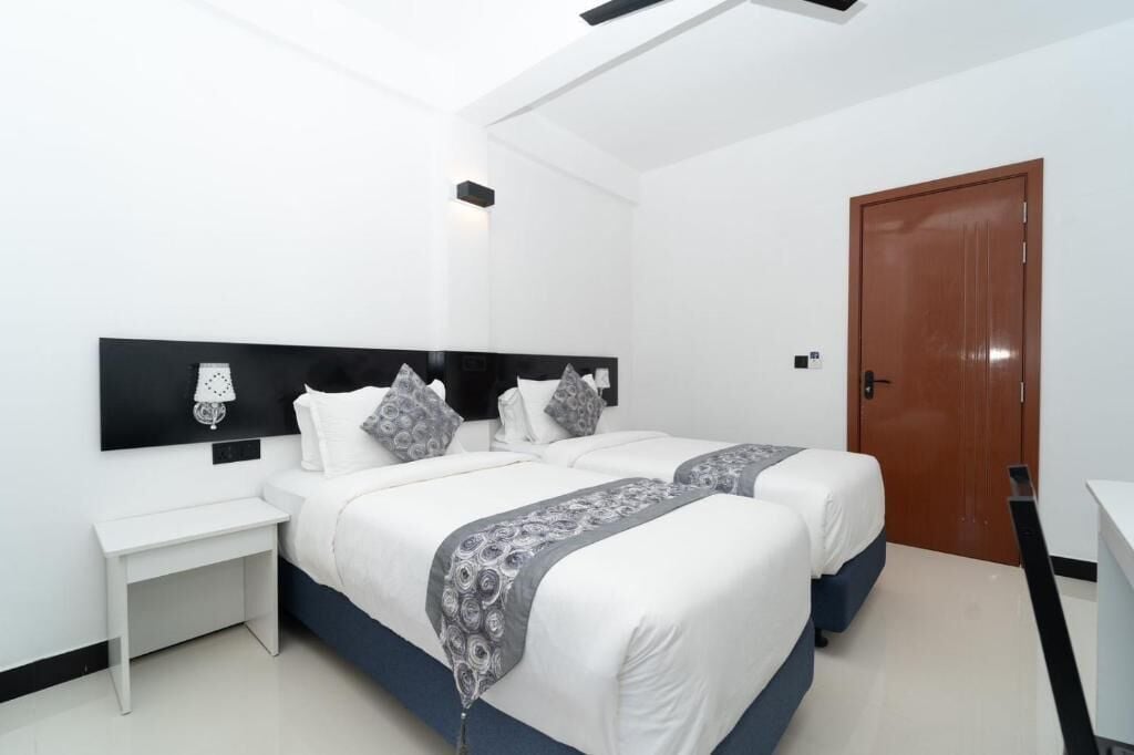 A modern hotel room with two beds, a white nightstand, a desk, and a wooden door.