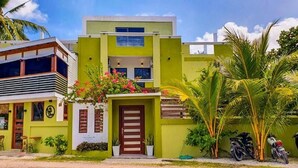 Un edificio moderno de dos pisos con fachada verde, una entrada central con puerta de madera y un pequeño jardín con macetas y flores.