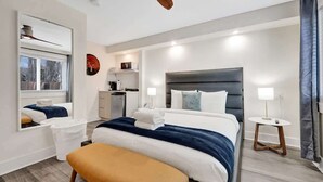 A modern hotel room with a large bed, a small desk, a mirror, a ceiling fan, and a window with blinds. - Chic Studio Steps to Fort Lauderdale Beach! (Fort Lauderdale)