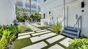 En modern byggnad med en stilren, geometrisk design, med en central gångväg som har konstgräs och betongplattor, omgiven av frodig grönska och en trappa som leder till en entré.