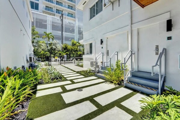 A modern building with a clean, geometric design, featuring a central pathway with artificial grass and concrete slabs, surrounded by lush greenery and a staircase leading to an entrance.