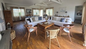 Een ruime woonkamer met een grote witte bank, twee rieten fauteuils, een houten salontafel en een keuken in de achtergrond.