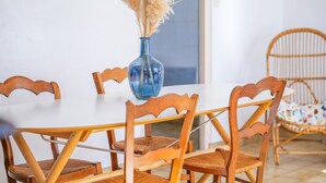 Una mesa de comedor con cuatro sillas de madera, un florero azul con plantas secas y una silla de mimbre al fondo.