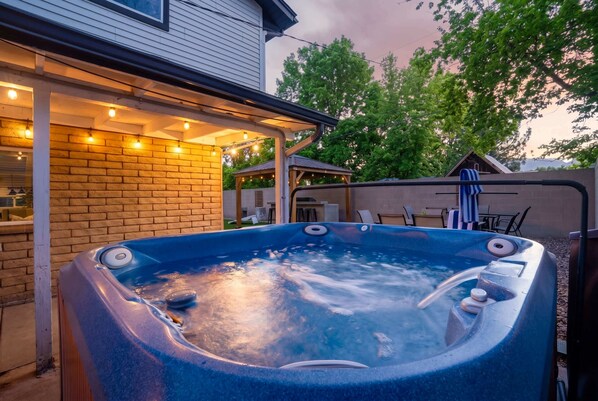 A hot tub in a backyard with a brick wall, outdoor lighting, and a covered patio area.