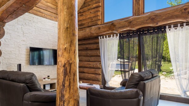 A log cabin interior with a stone wall, a flat-screen TV, leather sofas, and a view of the outdoors through sheer curtains. - Holiday Home Elizabeta (Slunj)