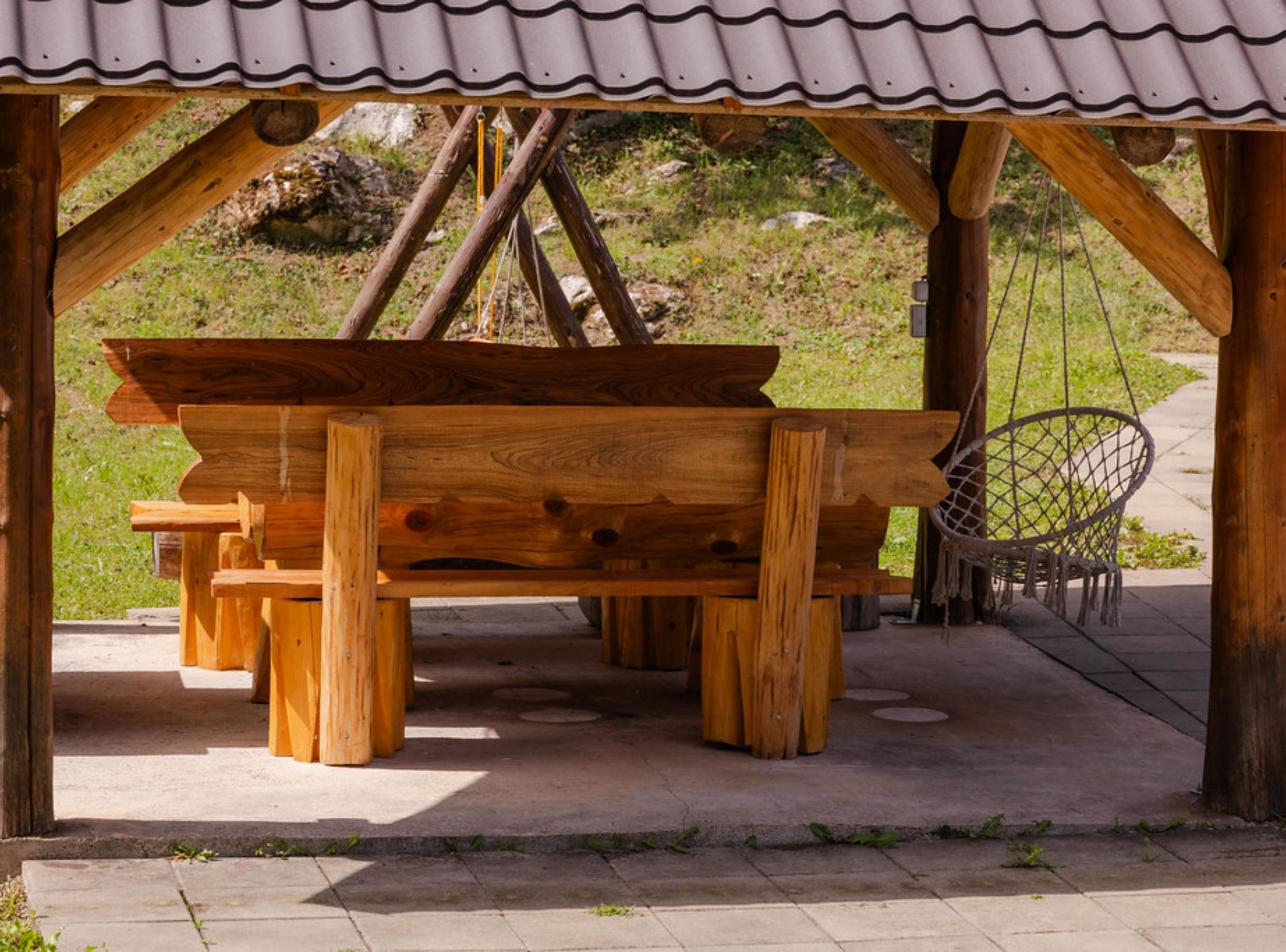 A wooden picnic table under a shelter with a corrugated roof, surrounded by a natural landscape.